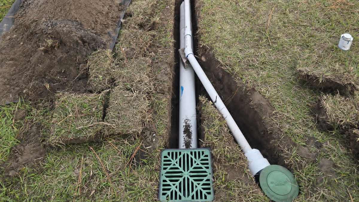 Open trench in a lawn with two white PVC downspout drain lines, one ending at a square green catch basin grate and one at a round green outlet