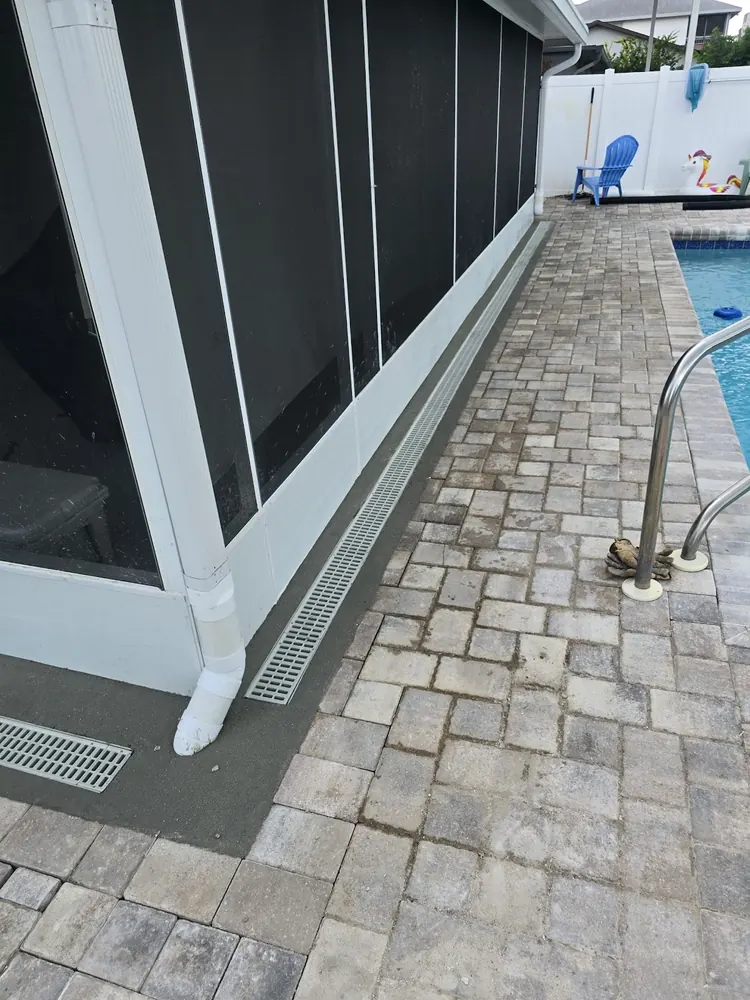 White downspout on a pool screen enclosure next to a long channel drain set into a paver pool deck