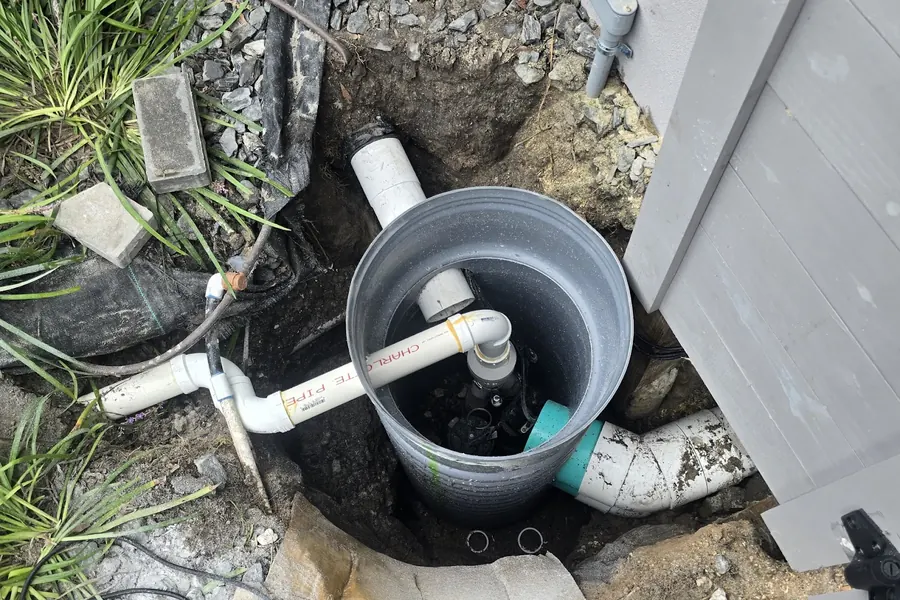Open round collection basin set in the ground beside a house with white PVC pipes connected to it