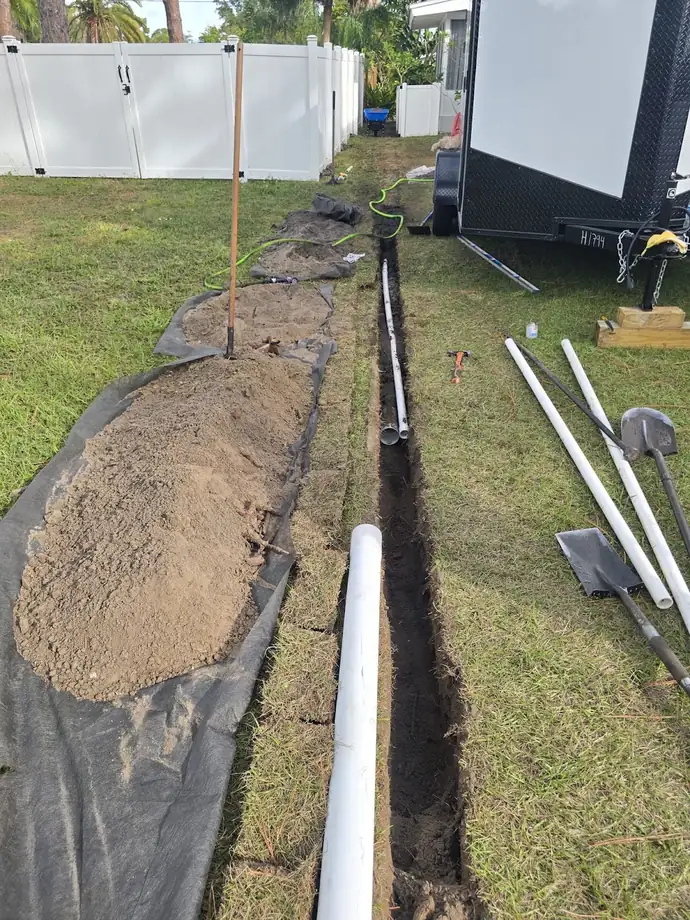 Narrow trench dug across a lawn with white PVC drain pipe laid in it and the soil piled on landscape fabric